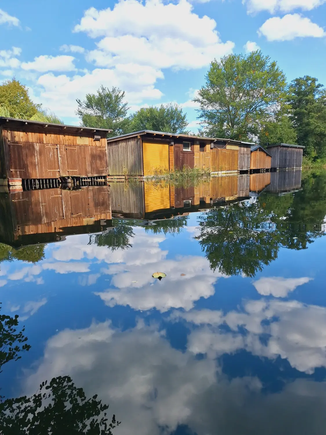Eine Reihe von hölzernen Bootshäusern am Rande eines ruhigen Gewässers, wahrscheinlich eines Sees oder Flusses.